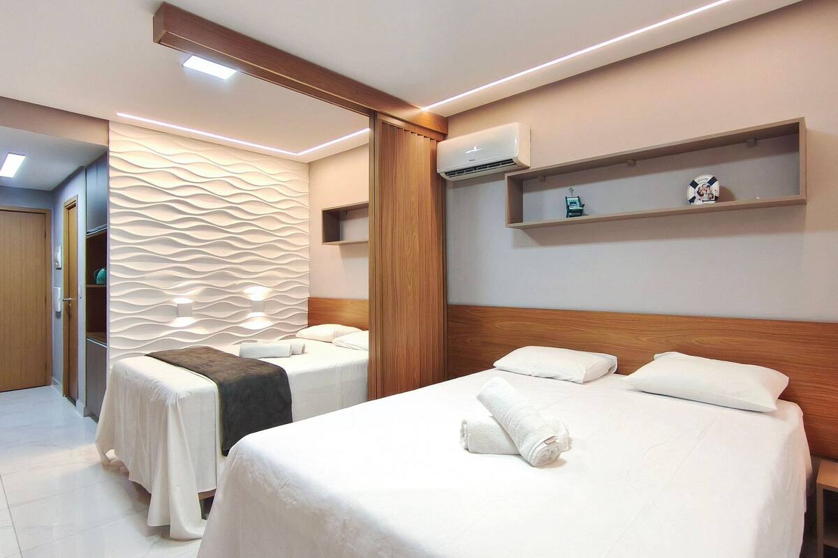 Habitación de hotel con dos camas, zona de masajes, cabecero de madera, equipo de aire acondicionado y mesita de noche con lámpara.
