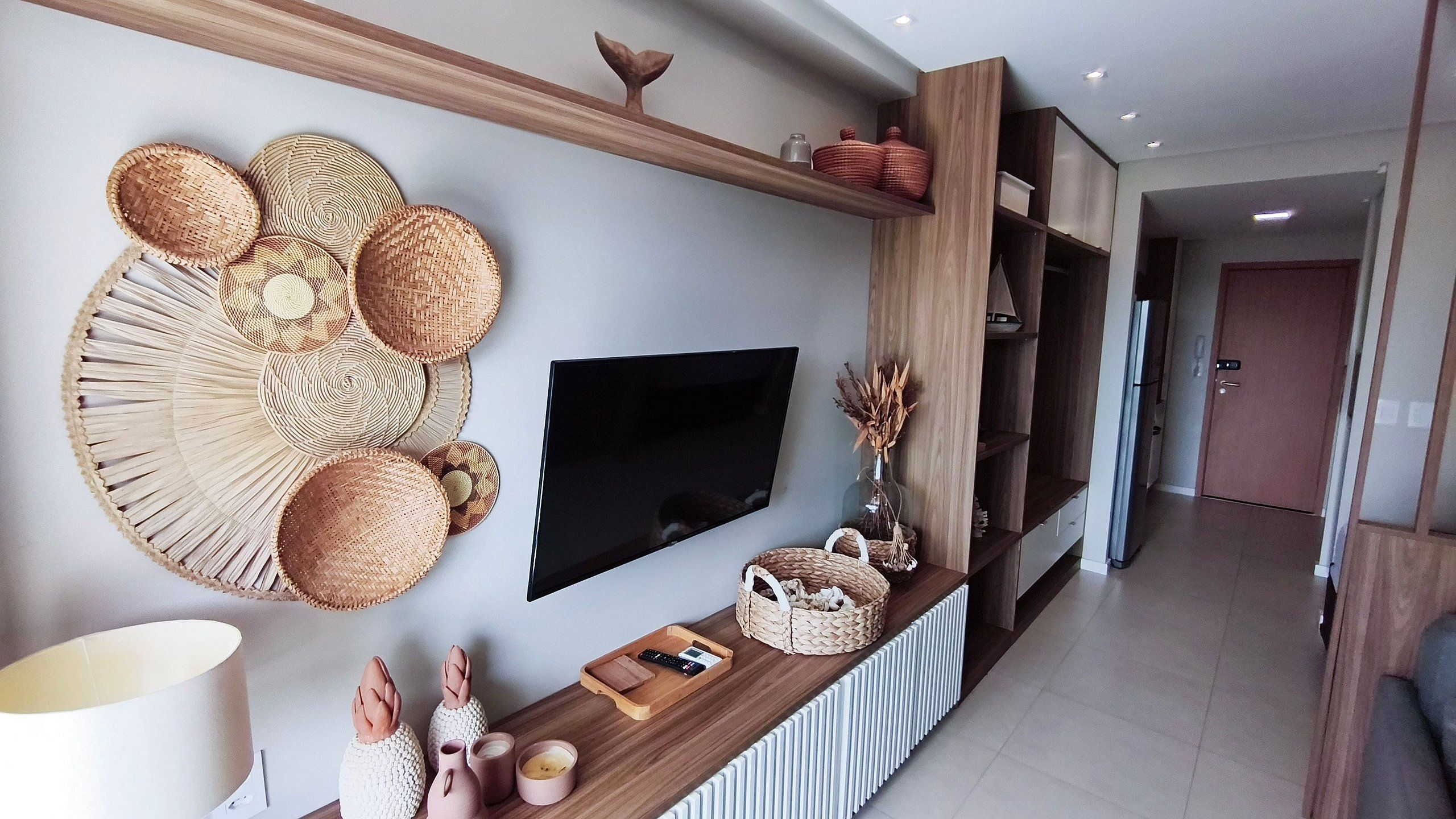 Un salon moderne doté d’un téléviseur à écran plat, d’une décoration murale en fibres tressées, d’une console en bois et d’un couloir menant à une porte.