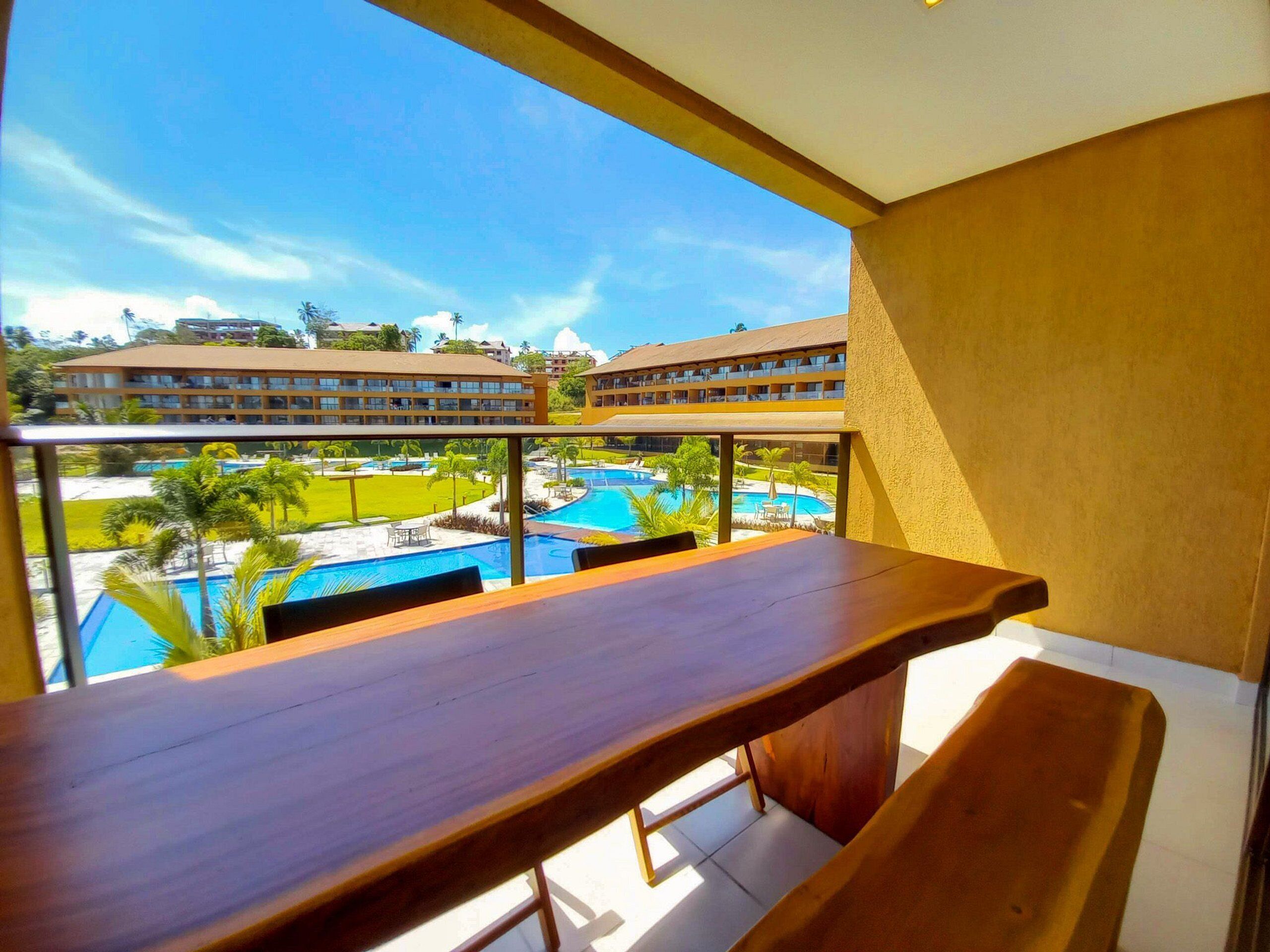 A balcony with a wooden table and bench, overlooking a pool and hotel buildings.