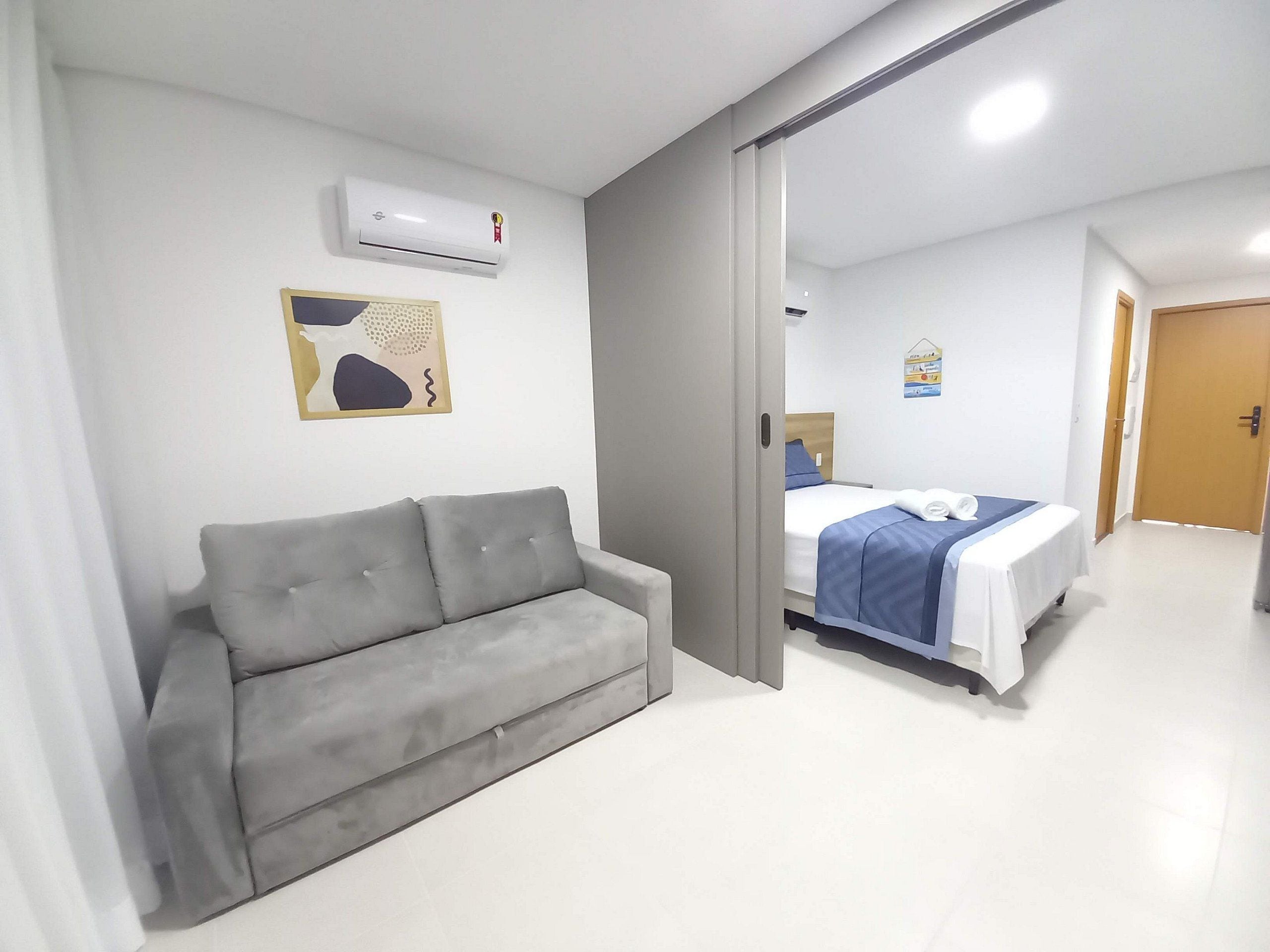 Ein Hotelzimmer mit grauem Sofa, einem Bett mit blauer Bettdecke, einer Klimaanlage und einem an der Wand befestigten Kunstwerk.