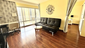 A living room with a black leather sofa, a small wooden coffee table, a desk with a chair, and a large window offering a view of high-rise buildings. - Waikiki Condo - Ocean View - Minutes from the Beach! (Honolulu)