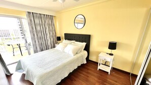 A hotel room with a bed, a bedside table with a lamp, a mirror on the wall, and a view of the city through the window. - Waikiki Condo - Ocean View - Minutes from the Beach! (Honolulu)