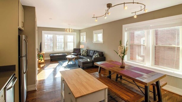 A modern kitchen with a built-in refrigerator, a dining area with a wooden table and benches, a gray sofa, and large windows providing natural light. - Chic Minimalist Gem w/ Fenced Backyard + W/D (Saint Louis)