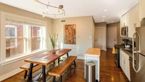 A modern kitchen with wooden flooring, a dining table with benches, a hanging light fixture, and a view of a brick building through large windows. - Chic Minimalist Gem w/ Fenced Backyard + W/D (Saint Louis)
