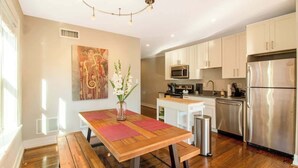 A modern kitchen with stainless steel appliances, a wooden dining table with a vase of flowers, and a painting on the wall. - Chic Minimalist Gem w/ Fenced Backyard + W/D (Saint Louis)