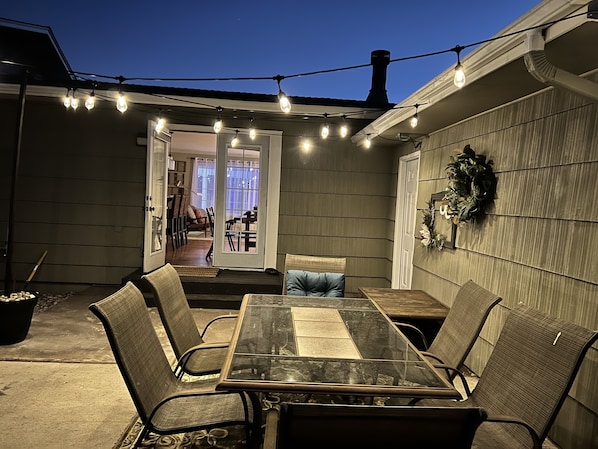 En udendørs terrasse med et bord og stole med glasplade, lyskæder hængt op og udsigt til huset.