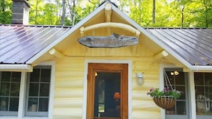 A log cabin with a wooden door, a hanging sign above the door, and a hanging flower basket. - Cottage Overlooking Paradise Lake (Carp Lake)