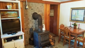 A cozy living room with a TV on a white cabinet, a black wood-burning stove, a dining table with chairs, and a painting on the wall. - Cottage Overlooking Paradise Lake (Carp Lake)