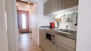 Una cocina moderna con electrodomésticos de acero inoxidable, horno empotrado y microondas. Hay un lavamanos con grifo y un hervidor sobre la encimera. La cocina tiene armarios de tonos claros y suelo de madera.