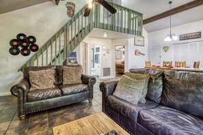 Un salon avec deux canapés en cuir, une table basse en bois, un escalier avec des rampes vertes et un coin repas avec des chaises en bois.