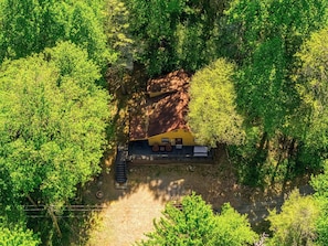Luchtbeeld van een gele hut met een onverharde pad er naartoe, omgeven door een dicht groen bos.