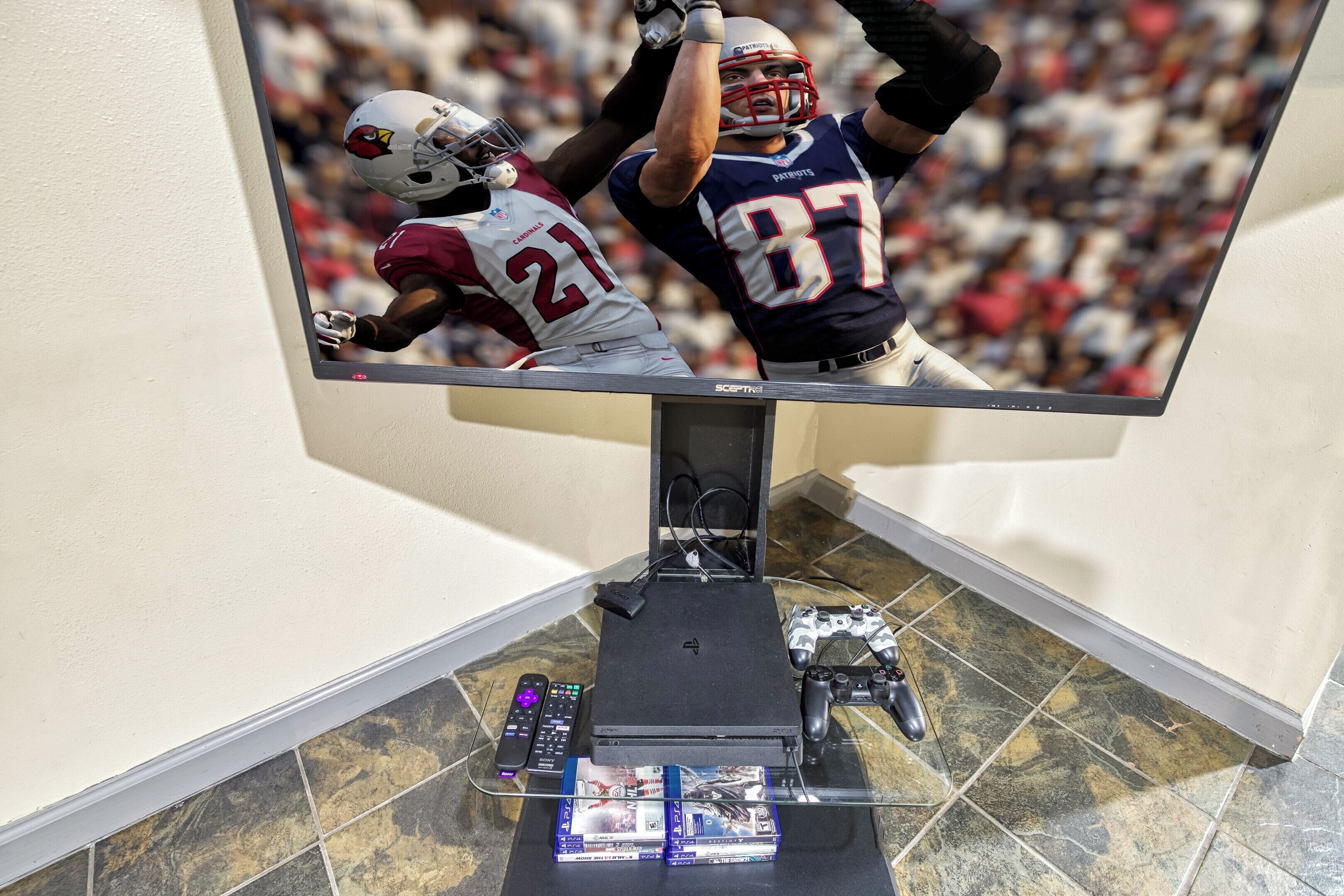Un televisor montado en la pared, con un control remoto, una consola PlayStation 4 y un mando de juegos sobre un pedestal.