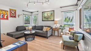 Un salon moderne avec un canapé d’angle gris, une table basse ronde et un fauteuil à motifs. Il y a deux fenêtres donnant sur la verdure, un luminaire au plafond et des œuvres d’art accrochées aux murs.
