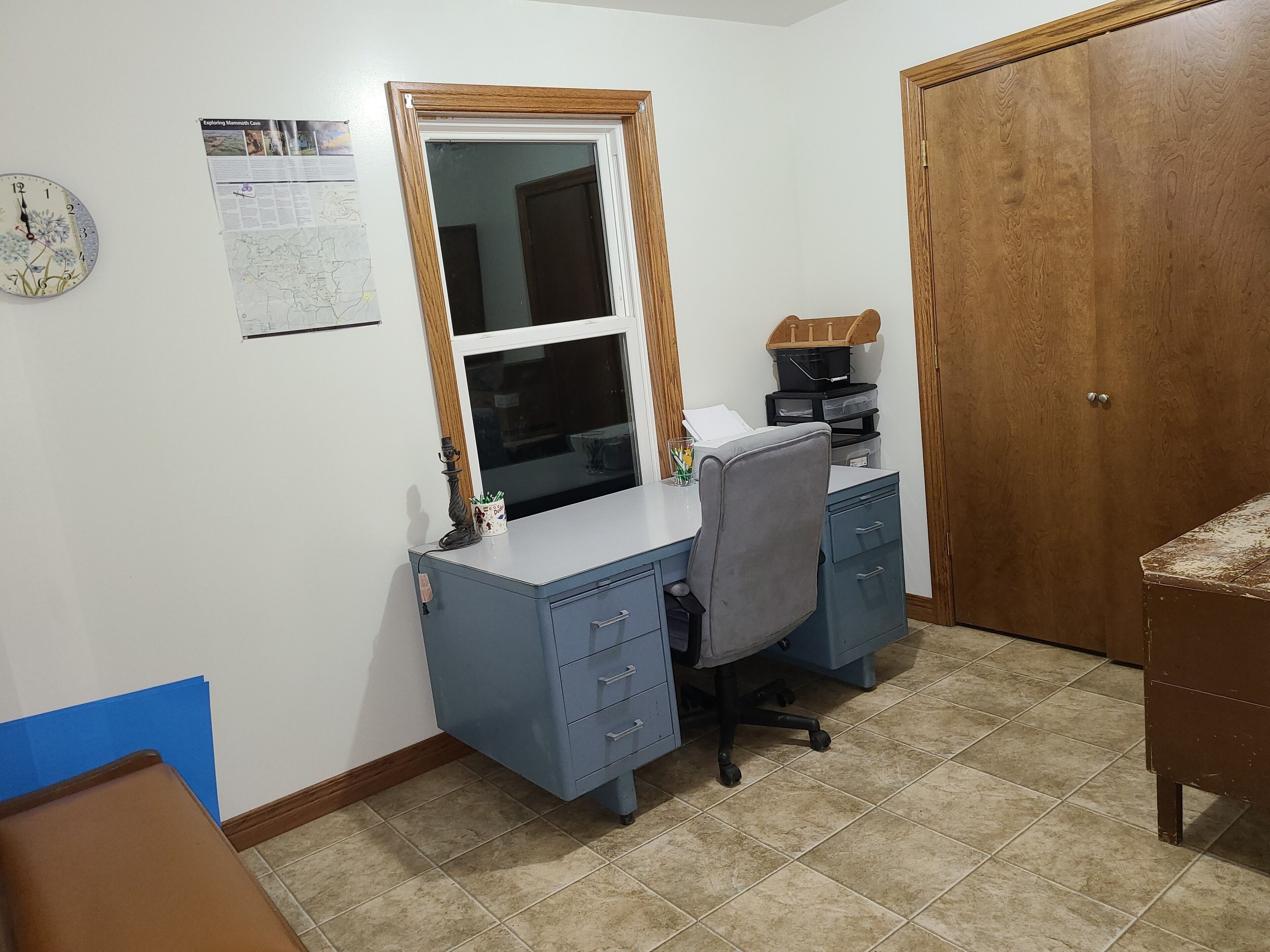 A small office with a desk, chair, and filing cabinet. There is a clock on the wall and a door to the right.
