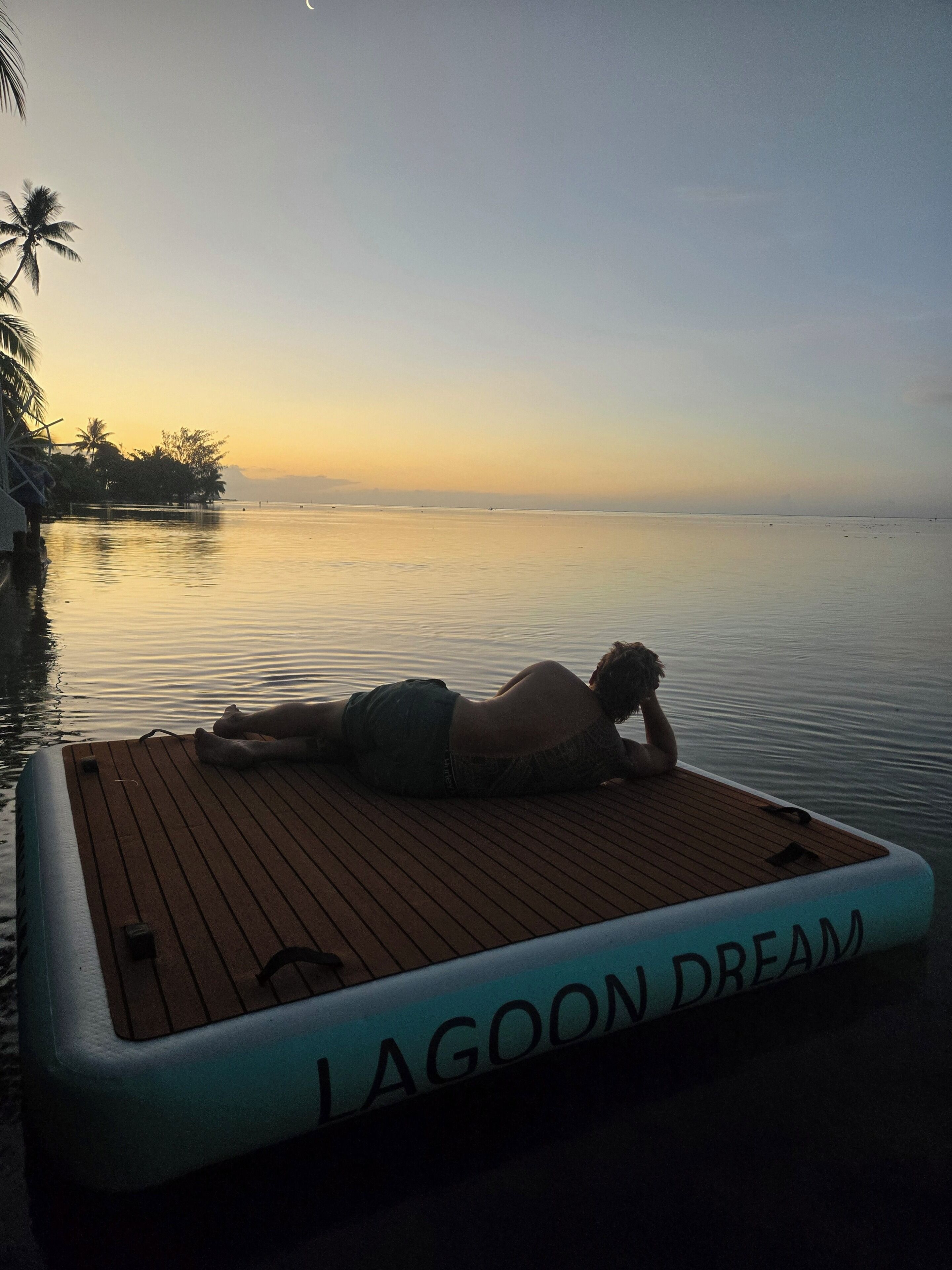 Una persona recostada sobre una cubierta de madera con la inscripción “LAGOON DREAM”, contemplando un agua tranquila y palmeras.