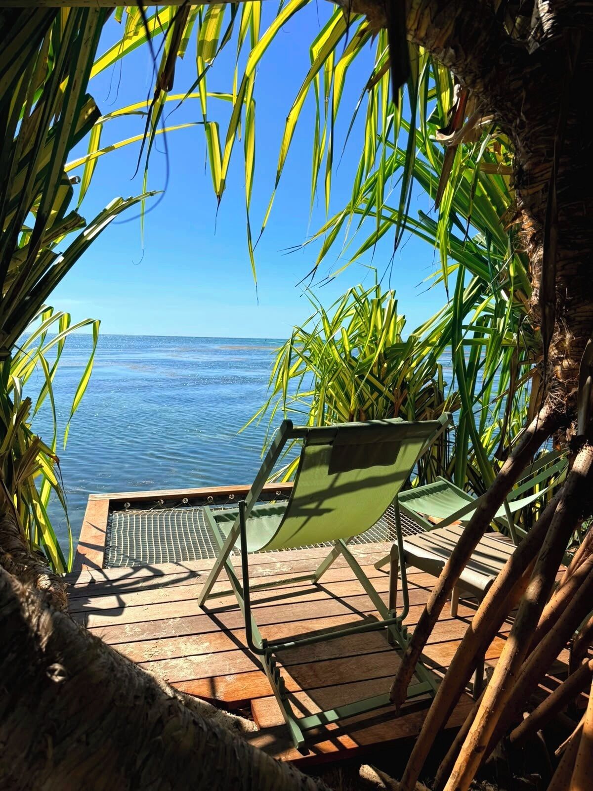 A wooden deck with a green chair and a small table, overlooking a clear blue sea.