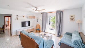 Ein Wohnzimmer mit einer Couch, einem Couchtisch, einem Fernseher, einem Deckenventilator und einem Fenster mit Vorhängen.