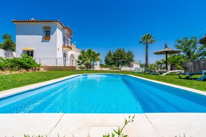 Une piscine avec un bâtiment blanc en arrière-plan, entourée de verdure et de palmiers.