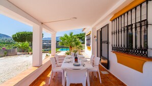 Eine überdachte Terrasse mit Esstisch für vier Personen, umgeben von weißen Stühlen, mit Blick auf einen Pool und einen Garten.