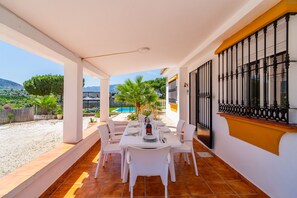 A covered patio with a dining table set for four, surrounded by white chairs, overlooking a swimming pool and garden.