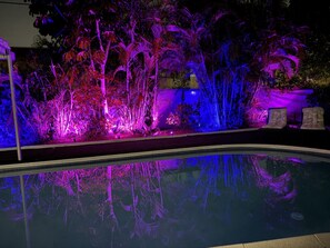 Ein Swimmingpool mit Spiegelung der umliegenden Vegetation und einer beleuchteten Landschaft bei Nacht.