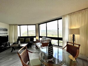 Relax in this stylish living room with a glass-top dining table, comfortable wicker chairs, and a sofa, offering a view of the city and hills.