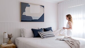 Een net opgemaakt bed met een kussen in blauw-witte patronen, een bedlampje en een lijst met een abstract schilderij aan de muur.