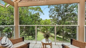 Een balkon met rieten meubels en een glazen tafel, met uitzicht op een tuin met diverse bomen en een heldere lucht.