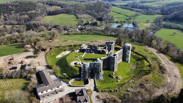 A historic castle with ruins, surrounded by green fields and a small building with a paved driveway.