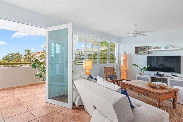 A modern living room with a sofa, a wooden coffee table, a TV, and a glass door leading to a balcony with a view of palm trees and buildings.