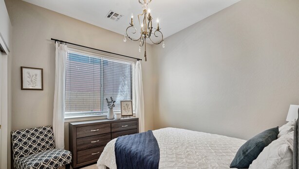 Une chambre à coucher comprenant un lit, une commode, une chaise, une fenêtre avec des stores et un lustre.