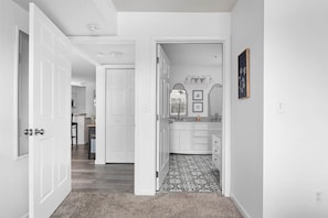 A hallway leading to a bathroom with a patterned floor, white walls, and a door with a silver handle.