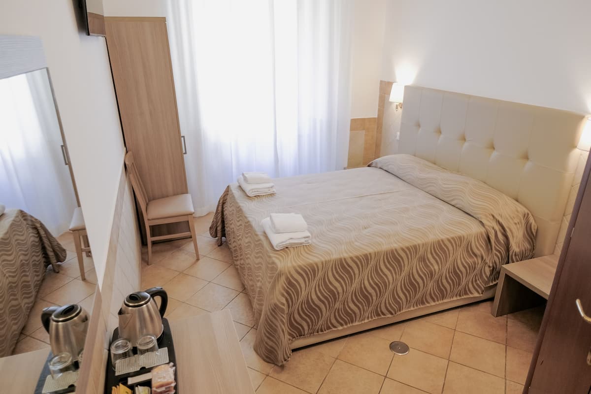 A hotel room with a bed, a chair, a desk with a kettle, cups, and a tray with food.