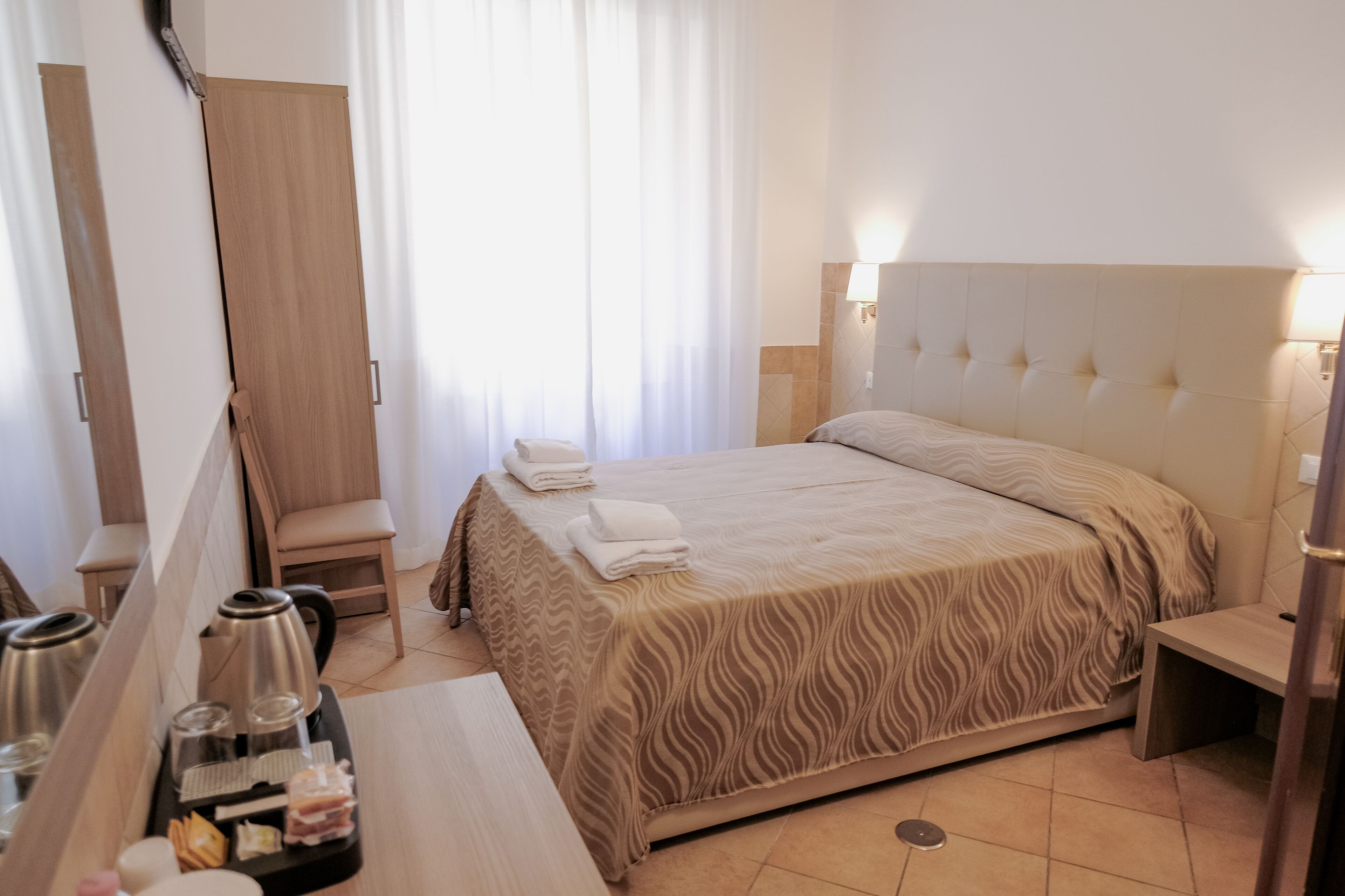 Une chambre d’hôtel avec un lit, une chaise, un bureau avec une bouilloire et un plateau de nourriture, et une fenêtre avec des rideaux.