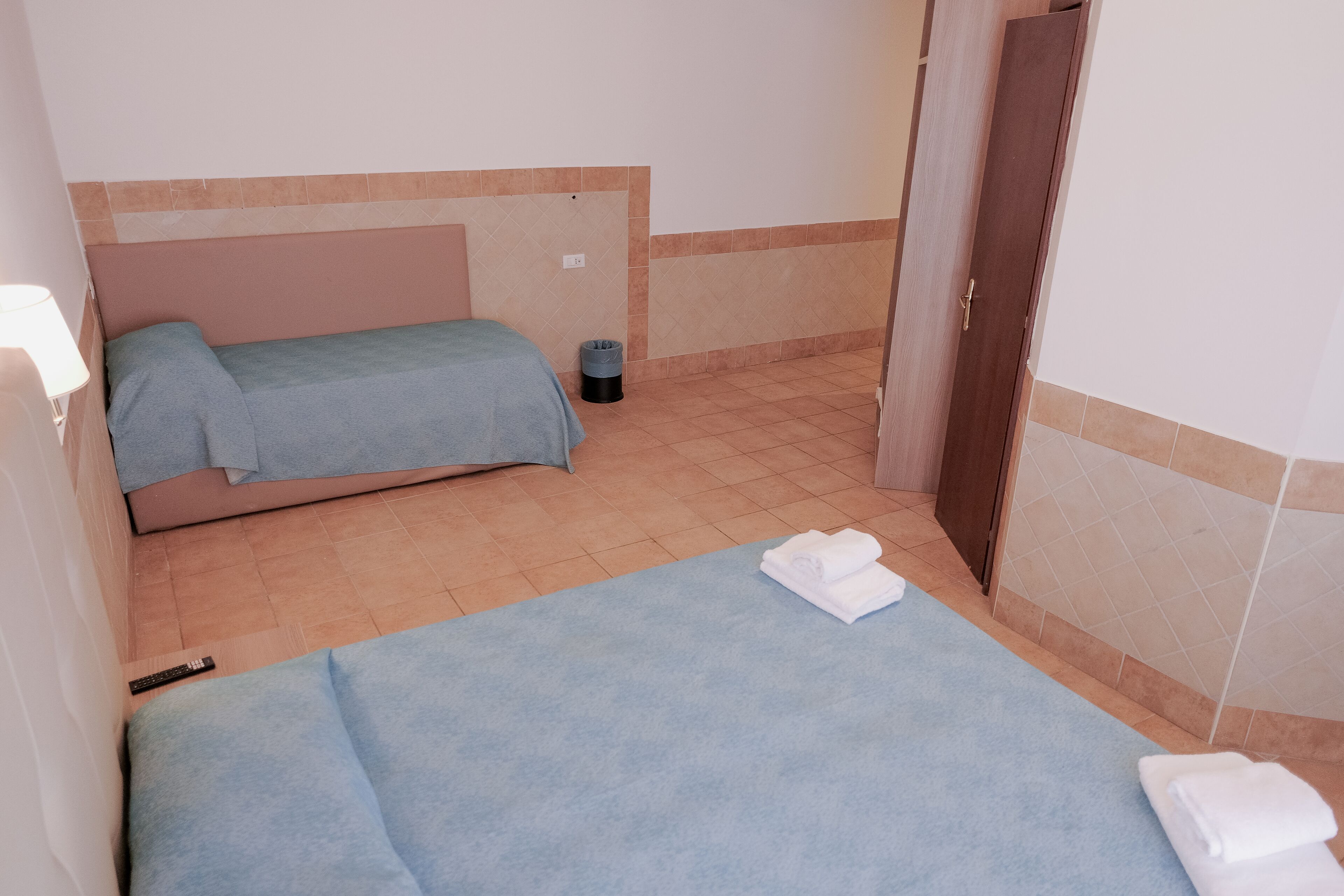 A small, single-bed room with a blue bedspread, a small trash bin, and a door leading to another room.