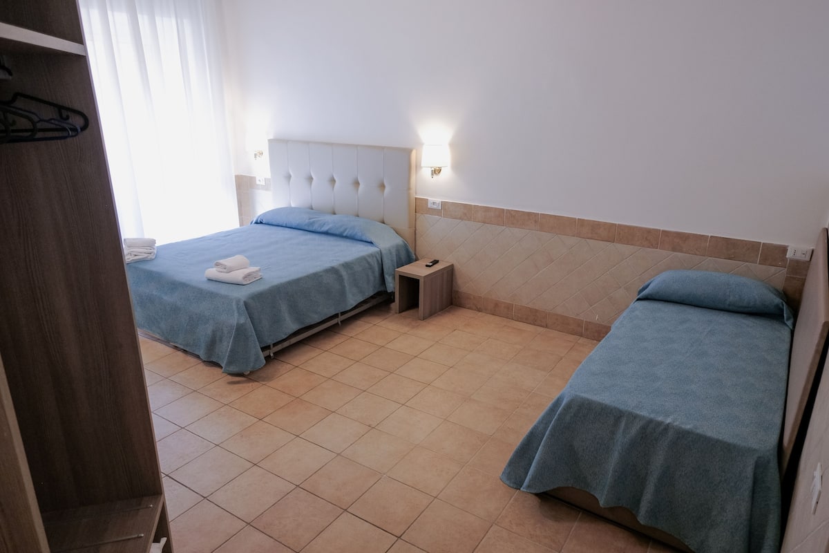 A hotel room with two single beds, a wooden wardrobe, a bedside table, and a wall-mounted light.
