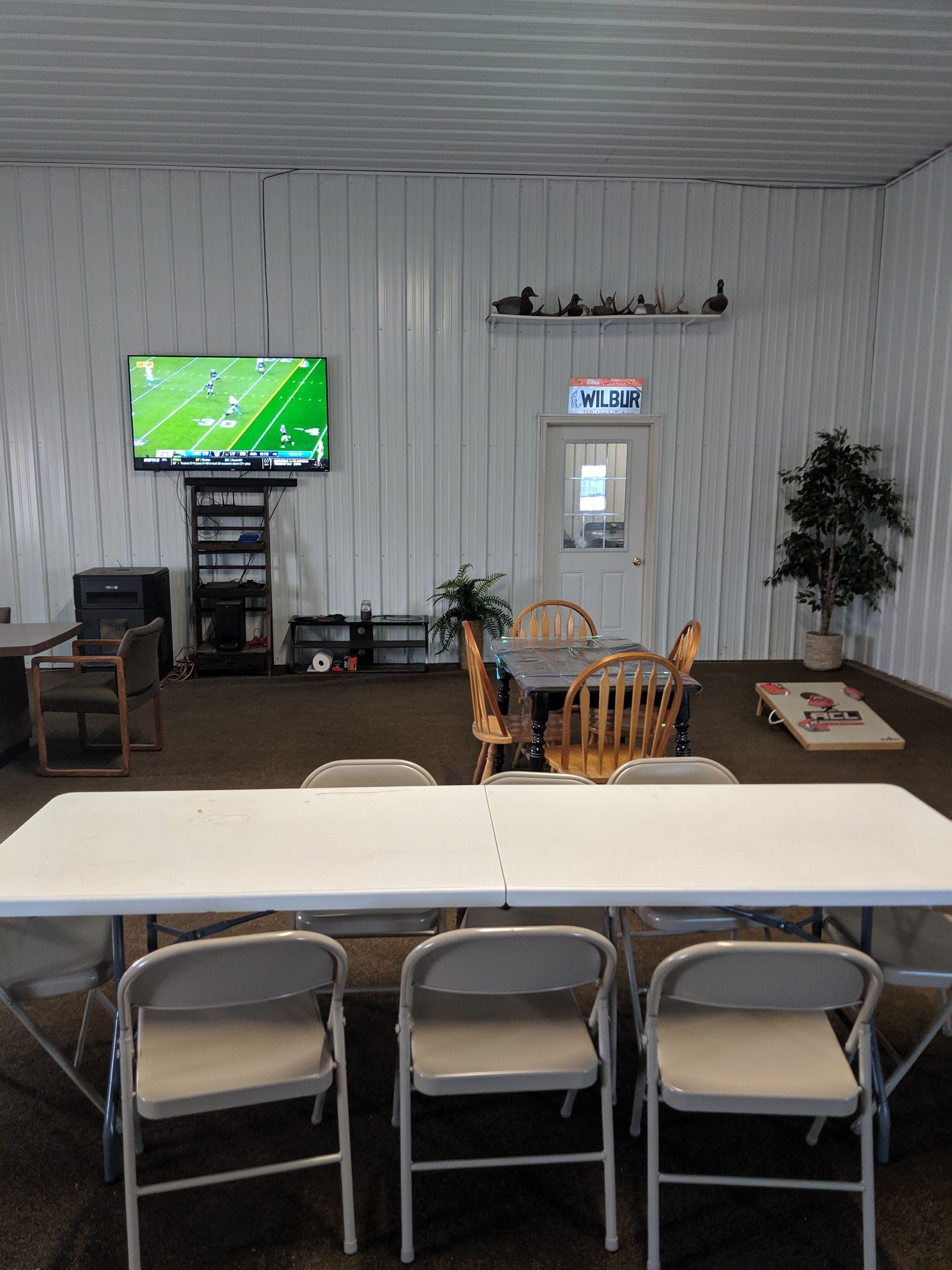 A room with a large flat-screen TV mounted on the wall, a dining table with chairs, a small plant, and a sign above the door.