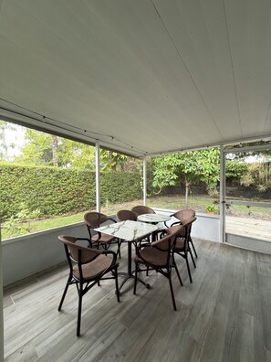 A covered patio with a table and chairs, large windows, and a view of greenery.