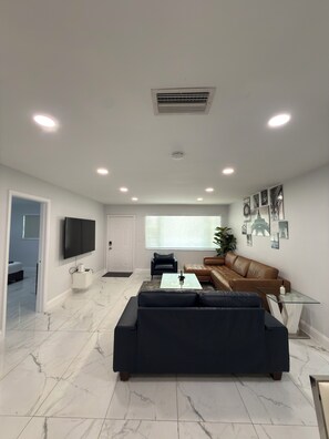 A modern living room with a large black sofa, a glass coffee table, a brown leather couch, and a flat-screen TV mounted on the wall.