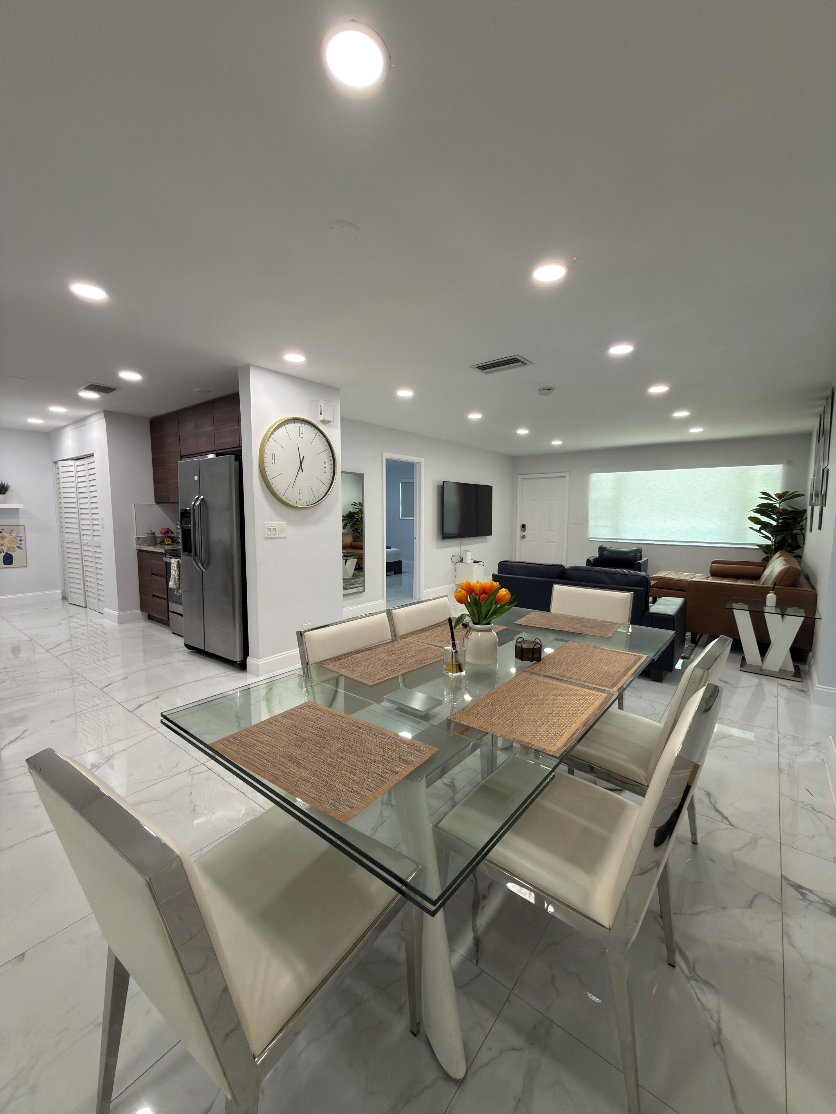 A modern kitchen with a refrigerator, a dining table with chairs, and a living area with a sofa and TV.