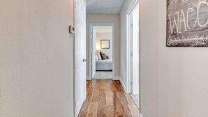 Ein Flur mit Holzboden und weißen Wänden, der zu einem Schlafzimmer mit Bett und einem gerahmten Bild führt.