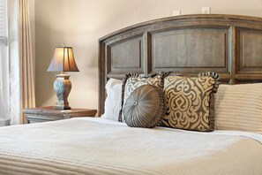 A neatly made bed with a white comforter and multiple decorative pillows, a bedside lamp, and a wooden headboard.