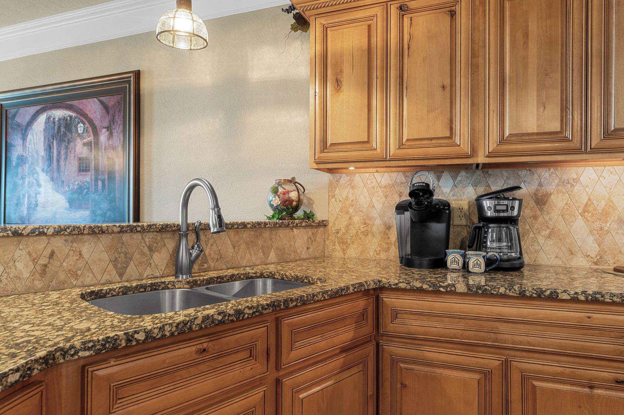 A kitchen with granite countertops, wooden cabinets, a stainless steel sink, a Keurig coffee maker, and a coffee pot.