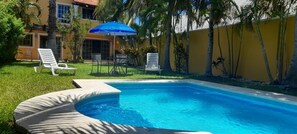 A swimming pool with lounge chairs and a blue umbrella, surrounded by palm trees and a yellow wall. - La Maison Bleue (Cancun)