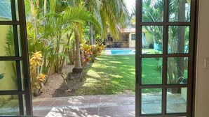 A view through a glass door into a backyard with a swimming pool, palm trees, and a grassy area. - La Maison Bleue (Cancun)