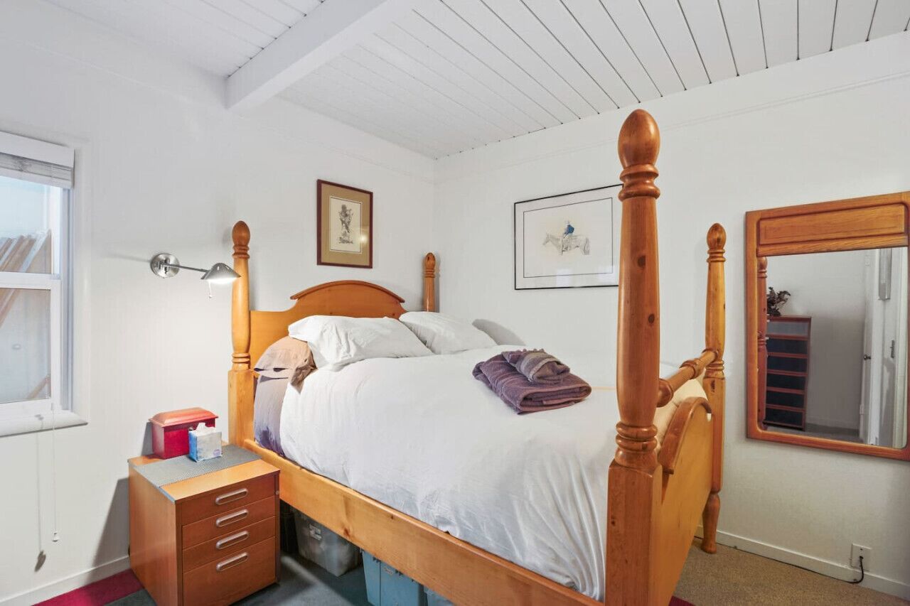 A bedroom with a wooden four-poster bed, a bedside table with drawers, a mirror, and framed artwork on the wall.
