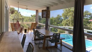 A spacious balcony with a wooden table and chairs, a hanging hammock, and a view of a swimming pool and trees. - Sea Cloud, Private home with salt water pool / Sea views (West Bay)