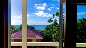 A view through a window of a building with a red roof, overlooking a body of water and a forested area. - Sea Cloud, Private home with salt water pool / Sea views (West Bay)