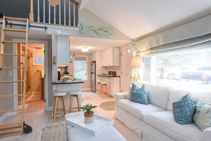 A modern living room with a white sofa, a coffee table with a plant, a ladder, and a kitchen area with white cabinets and stools.