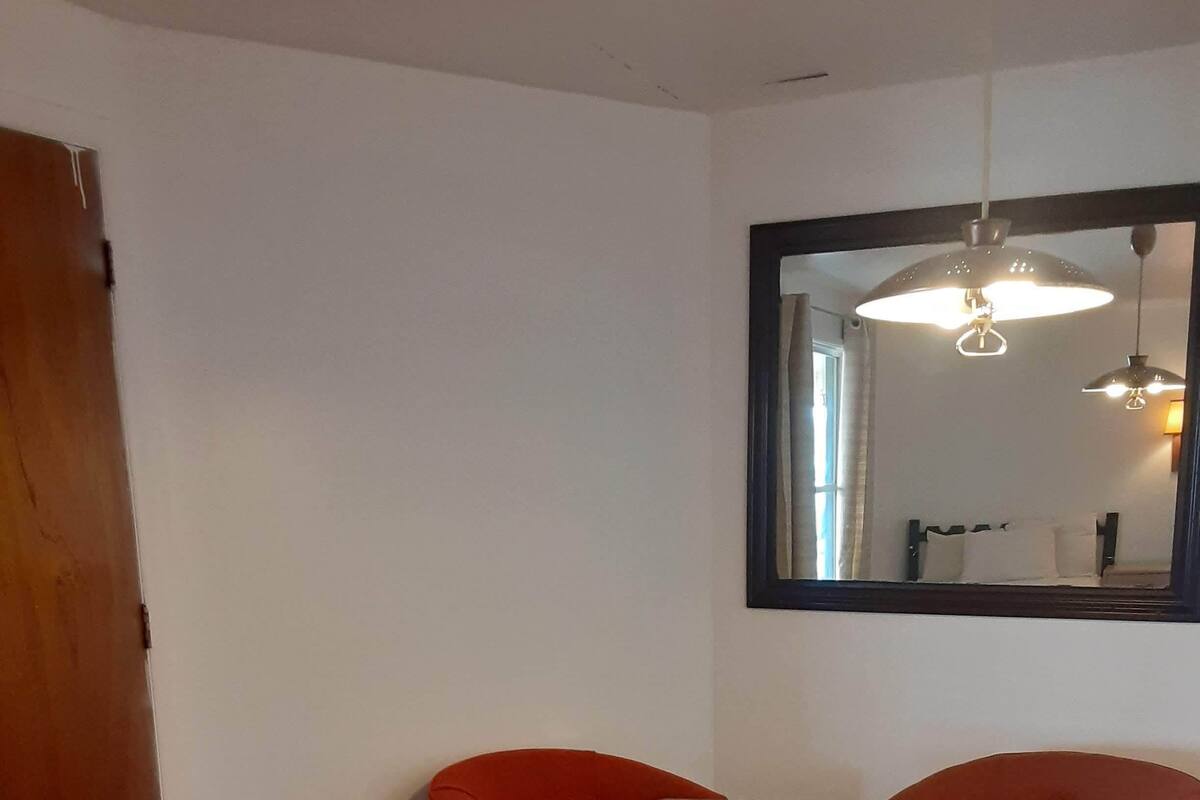 A room with a wooden floor, a small table, two red chairs, a wooden door, and a mirror on the wall.